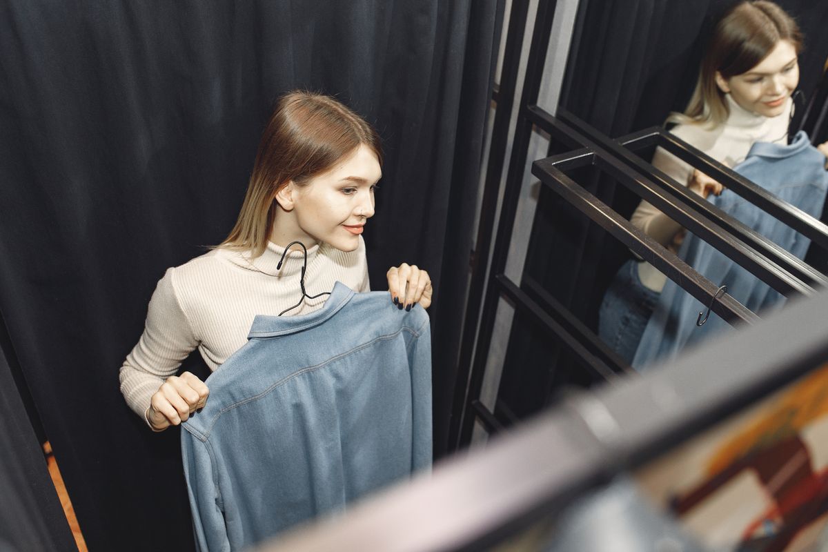 Woman in a fitting room holding a denim shirt beside a mirror, trying clothes on before buying