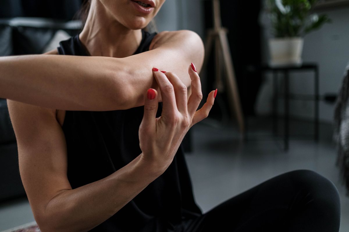 Woman in a dark sleeveless top with her arm raised across her chest, showing the front of the torso where a bra can creep upward